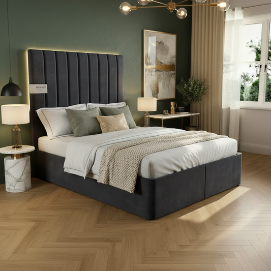 A light gray upholstered bed frame with a tall channel-tufted headboard and raised storage base, shown in a styled bedroom with wood flooring, a window, curtains, and a side table.