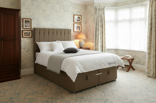 A bed with a brown upholstered headboard and matching base, dressed in white bedding, shown in a bright bedroom with wood flooring, a wall clock, a chair, and a small side table.