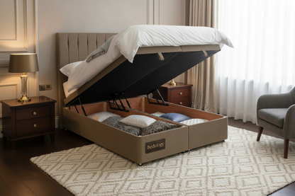 A lift-up storage bed frame in a warm taupe-brown fabric, shown with the mattress lifted open to reveal storage compartments underneath, set in a bright bedroom with wooden floors and a wooden chair.