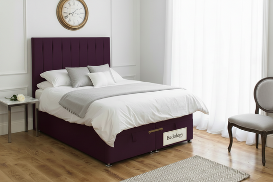 A double bed with a deep purple upholstered headboard and matching storage base, dressed in white bedding, set in a bright bedroom with wood floors, a wall clock, and a wooden chair.