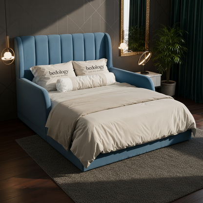 A blue upholstered bed with a tall channel-tufted headboard and matching footboard, styled in a bedroom with cream bedding, two pillows printed with the word 'bedology', a small side table with a lamp, and a potted plant.