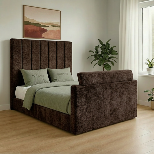 Brown upholstered bed with green bedding in a room with a window and plants.